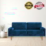 An Alia Velvet 3 Seater Square Arm Sofa in blue graces a minimalist living room with light wood flooring. To the left, two vases and a candle add coziness. A Premium Quality Guaranteed badge and a 5 Years Warranty badge on the wall invite you to shop for timeless style and lasting assurance.