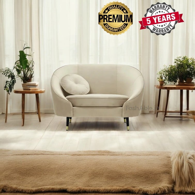 A stylish beige armchair with a round cushion sits in a bright room. Flanked by wooden side tables with potted plants, the space exudes charm. Shop now for this chic setting, where sheer curtains and a soft rug enhance the elegance, and quality badges assure your confidence in buying.