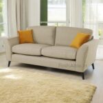 The Mahir Lenka Fabric 3+1+1 Seater Sofa Set, in beige with two mustard-yellow cushions, is stylishly placed in a well-lit room with large windows. A textured beige rug partially covers the wooden floor as sunlight creates soft shadows, enhancing the warm and modern design ambiance.